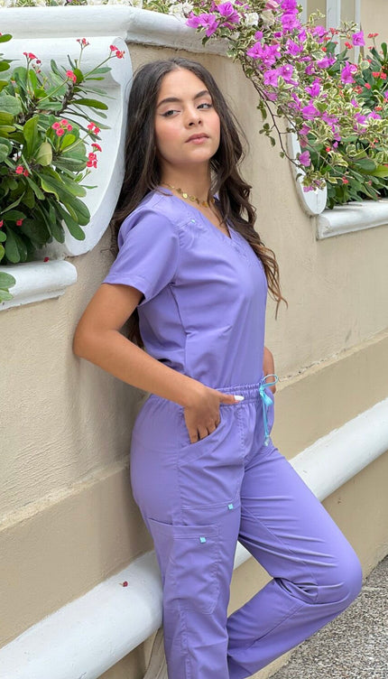 Person wearing a purple scrub set leaning against a wall with plants.