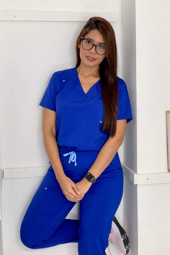 Woman wearing a blue scrub set sitting against a white wall.